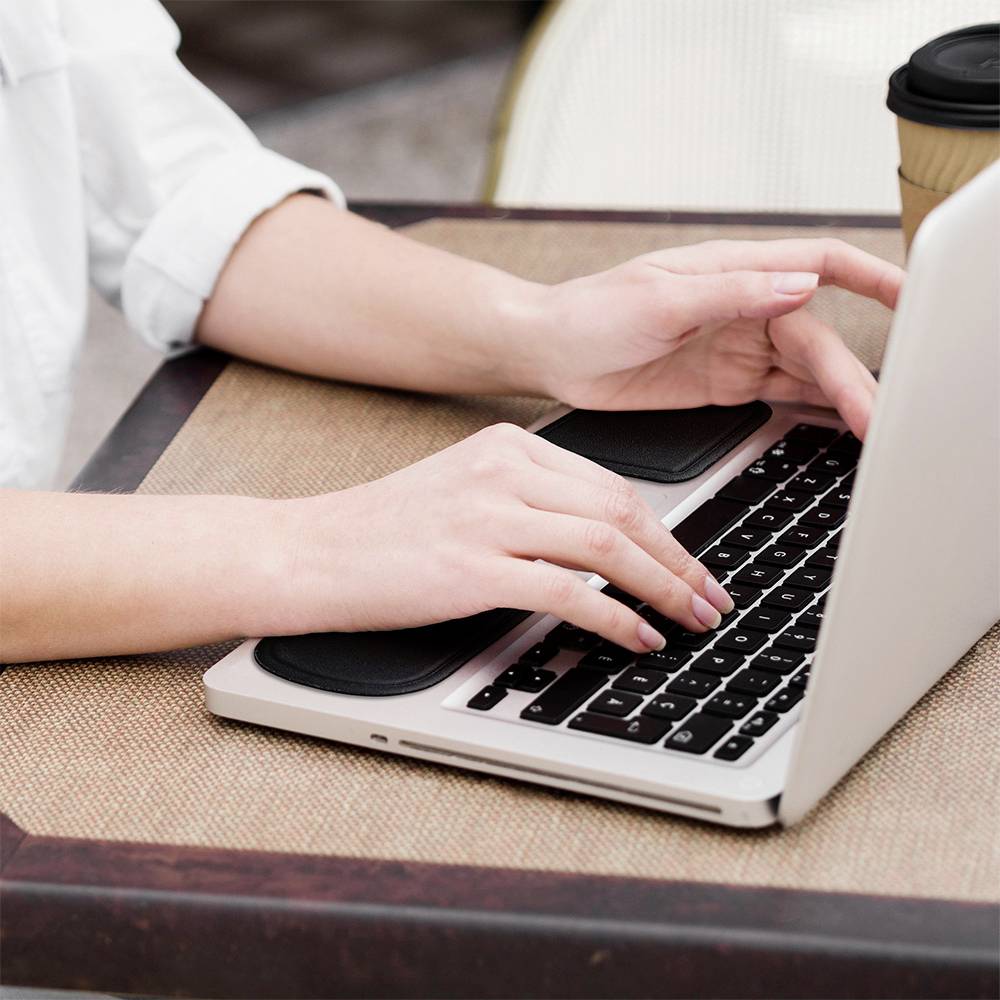 Eine Person tippt auf einem Laptop und hält einen Kaffeebecher. Sie sitzt an einem Tisch.