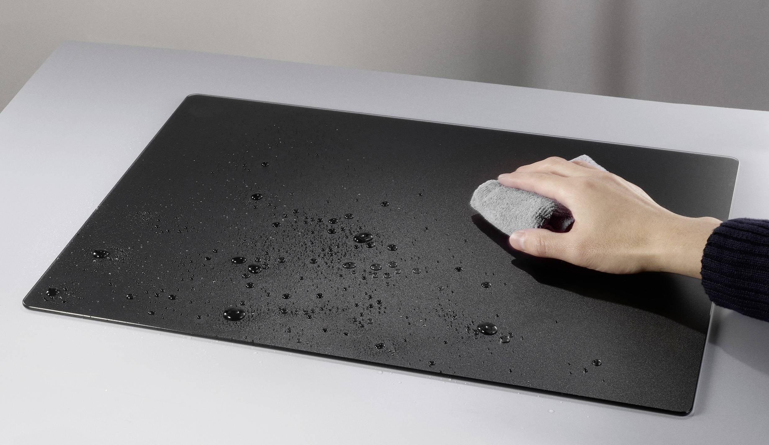 Eine Hand wischt Wassertropfen von einer schwarzen, rechteckigen Oberfläche auf einem grauen Tisch mit einem grauen Tuch ab.