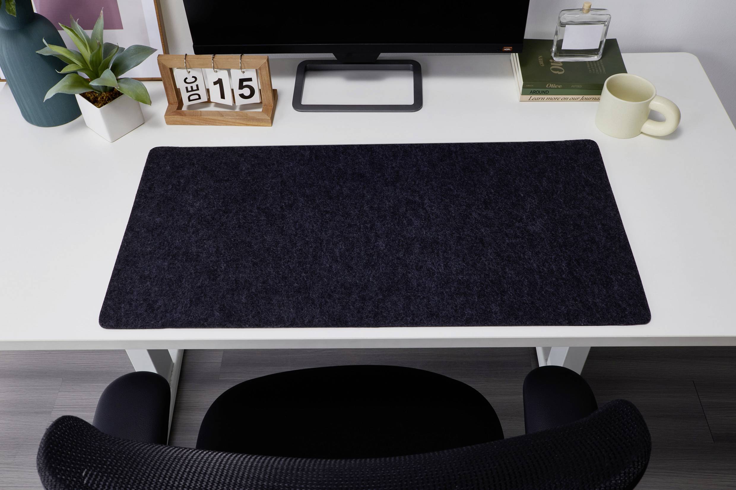 Schreibtisch mit schwarzer Schreibtischunterlage, Kalender zeigt '15 Dez', Monitor, Pflanze, Buch, Tasse und Brille daneben.