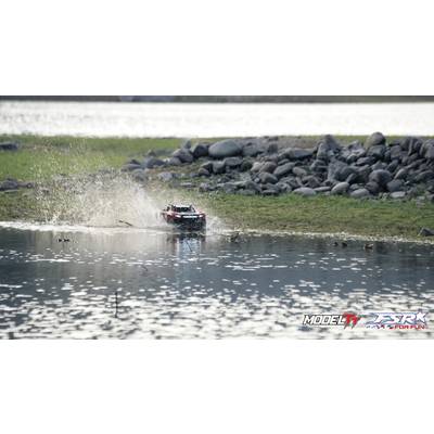 Ein ferngesteuertes Auto fährt durch flaches Wasser, spritzt Wasser auf, umgeben von Steinen und einem See im Hintergrund.