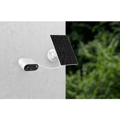Überwachungskamera mit Solarpanel an einer Wand befestigt, bei Regen in einem Außenbereich mit grüner Vegetation im Hintergrund.