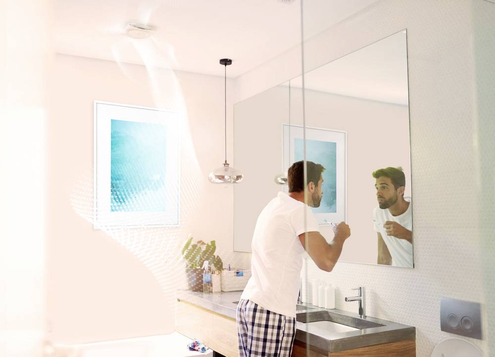 Ein Mann putzt sich die Zähne vor einem Badezimmerspiegel. Im Hintergrund sind ein Waschbecken, eine Lampe und ein Bild zu sehen.