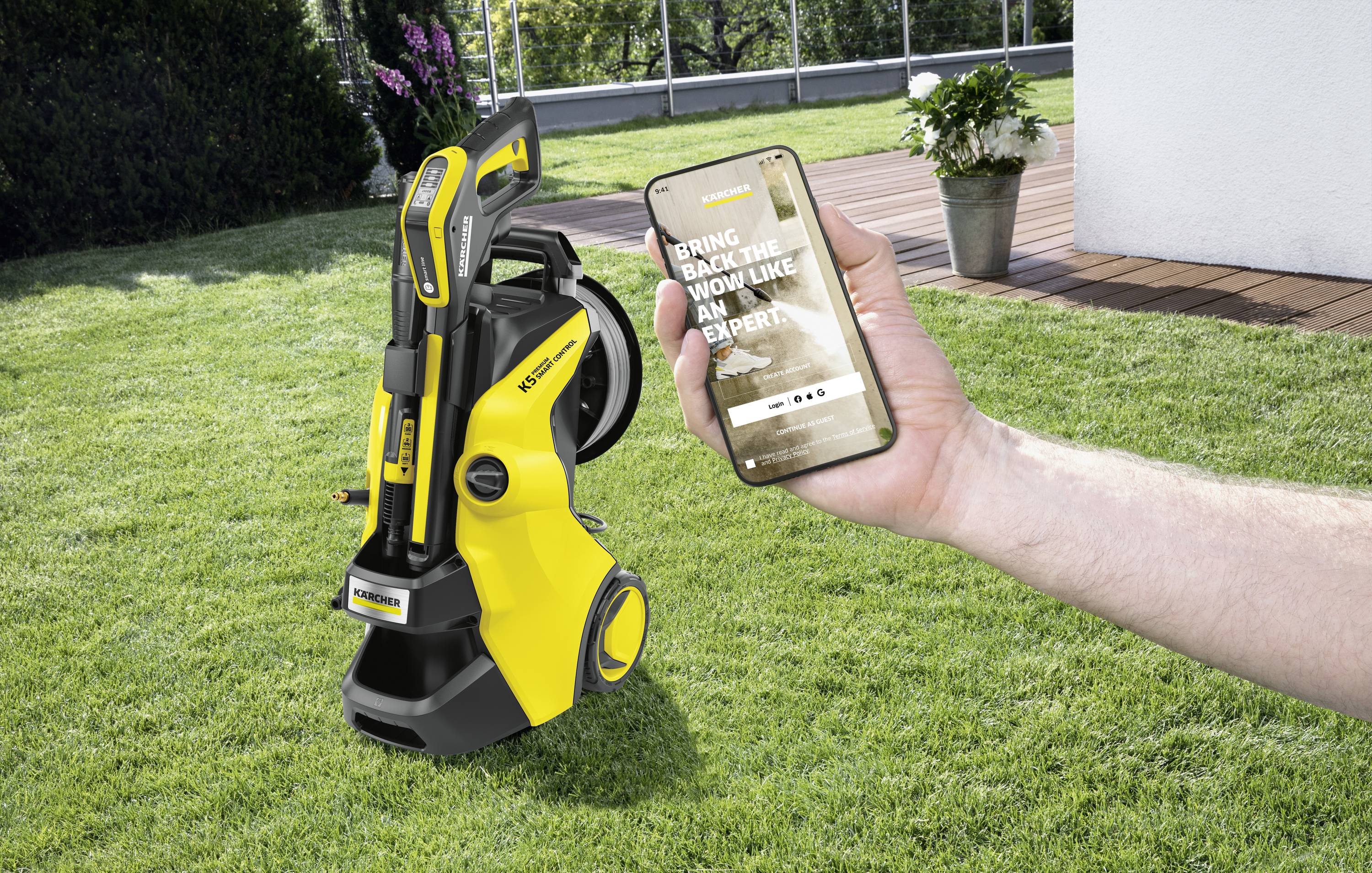 Eine Person hält ein Smartphone mit geöffneter App, während sie in einem Garten steht. Daneben steht ein gelber Hochdruckreiniger.