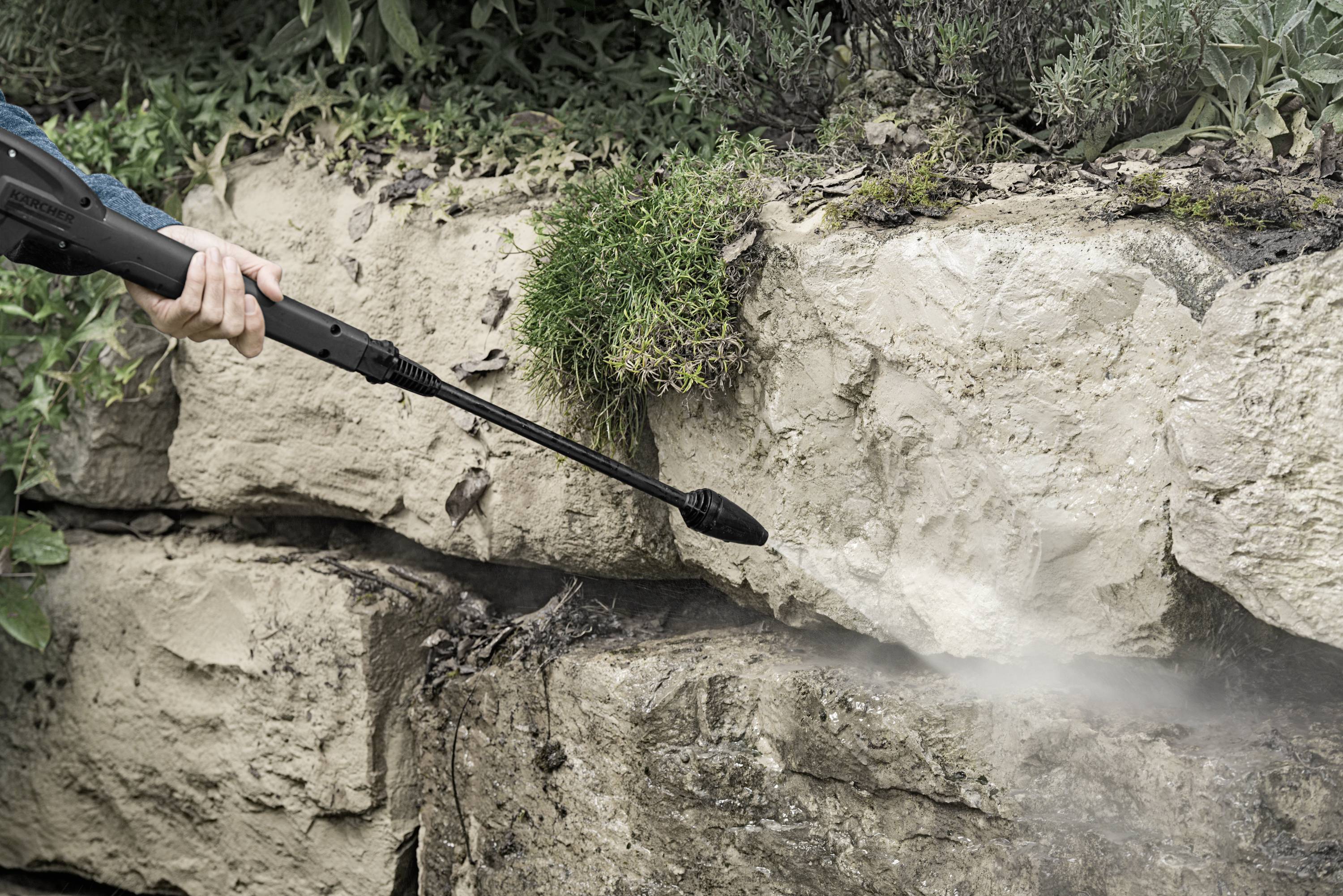 Eine Person reinigt mit einem Hochdruckreiniger eine steinerne Mauer. Wasser spritzt auf die Felsen und entfernt Schmutz.