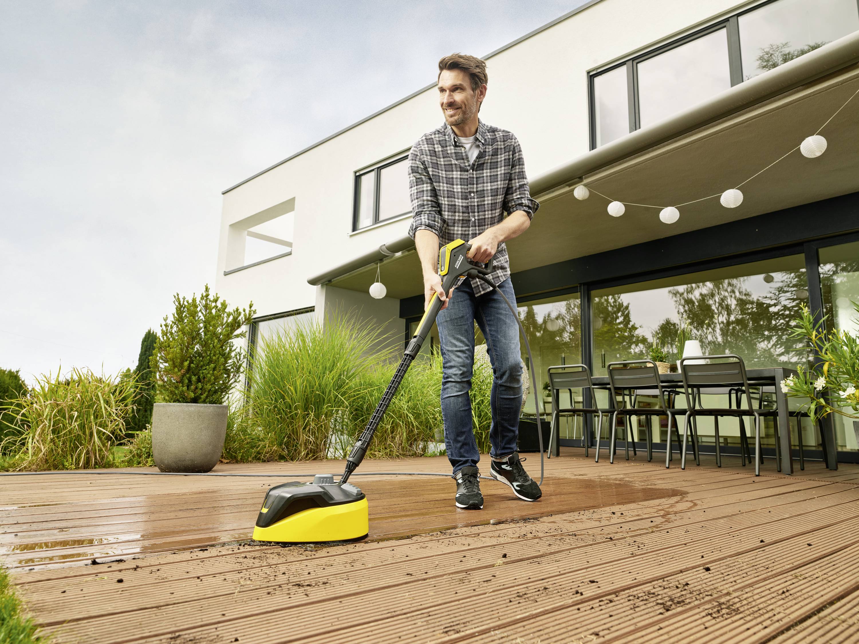 Ein Mann reinigt eine Holzterrasse mit einem Hochdruckreiniger vor einem modernen Haus, umgeben von Pflanzen und Gartenmöbeln.