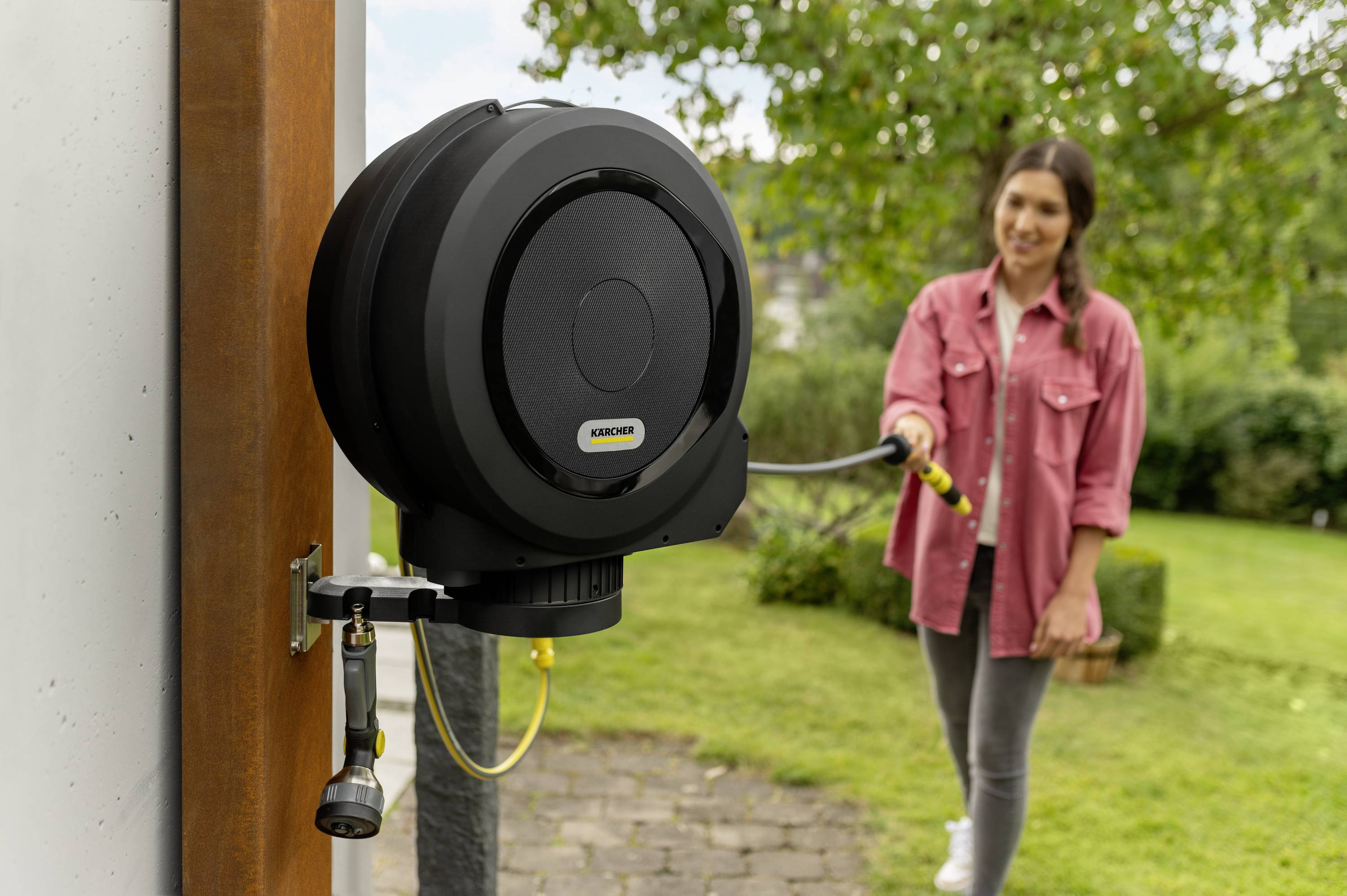 Frau im Garten zieht einen Gartenschlauch aus einer automatisch aufrollbaren Schlauchtrommel, befestigt an einer Wand.