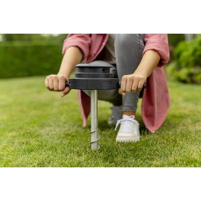 Person installiert ein Gerät im Rasen mit einem Handgriff und einer Schraube. Sie trägt Sneakers und eine rote Jacke. Im Hintergrund ist Garten zu sehen.