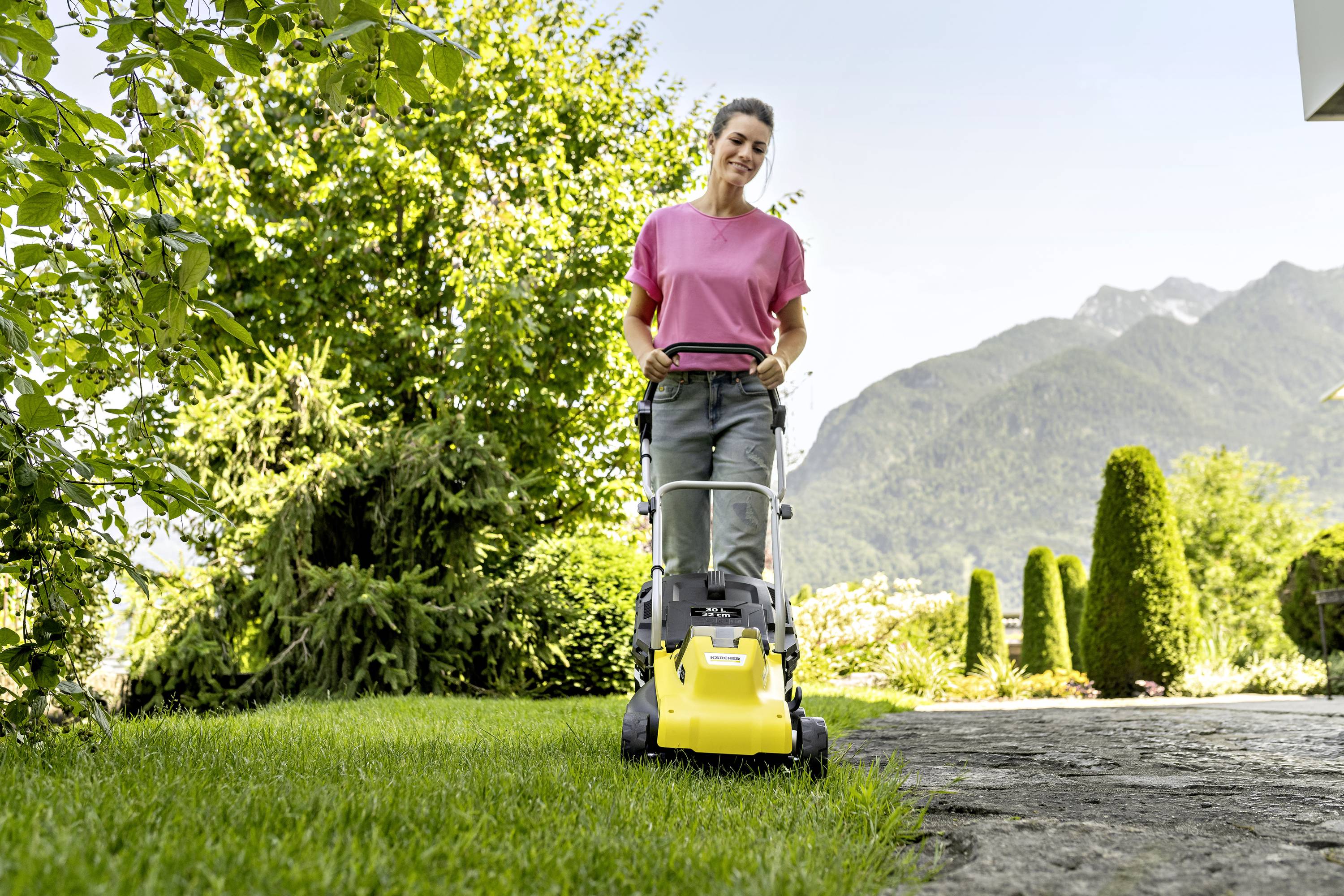 Eine Person mäht mit einem gelben Rasenmäher das Gras in einem sonnigen Garten. Im Hintergrund sind Berge sichtbar.