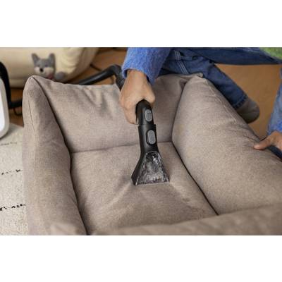 Eine Person reinigt ein braunes Hundebett mit einem Handstaubsauger. Im Hintergrund ist ein Hundespielzeug zu sehen.