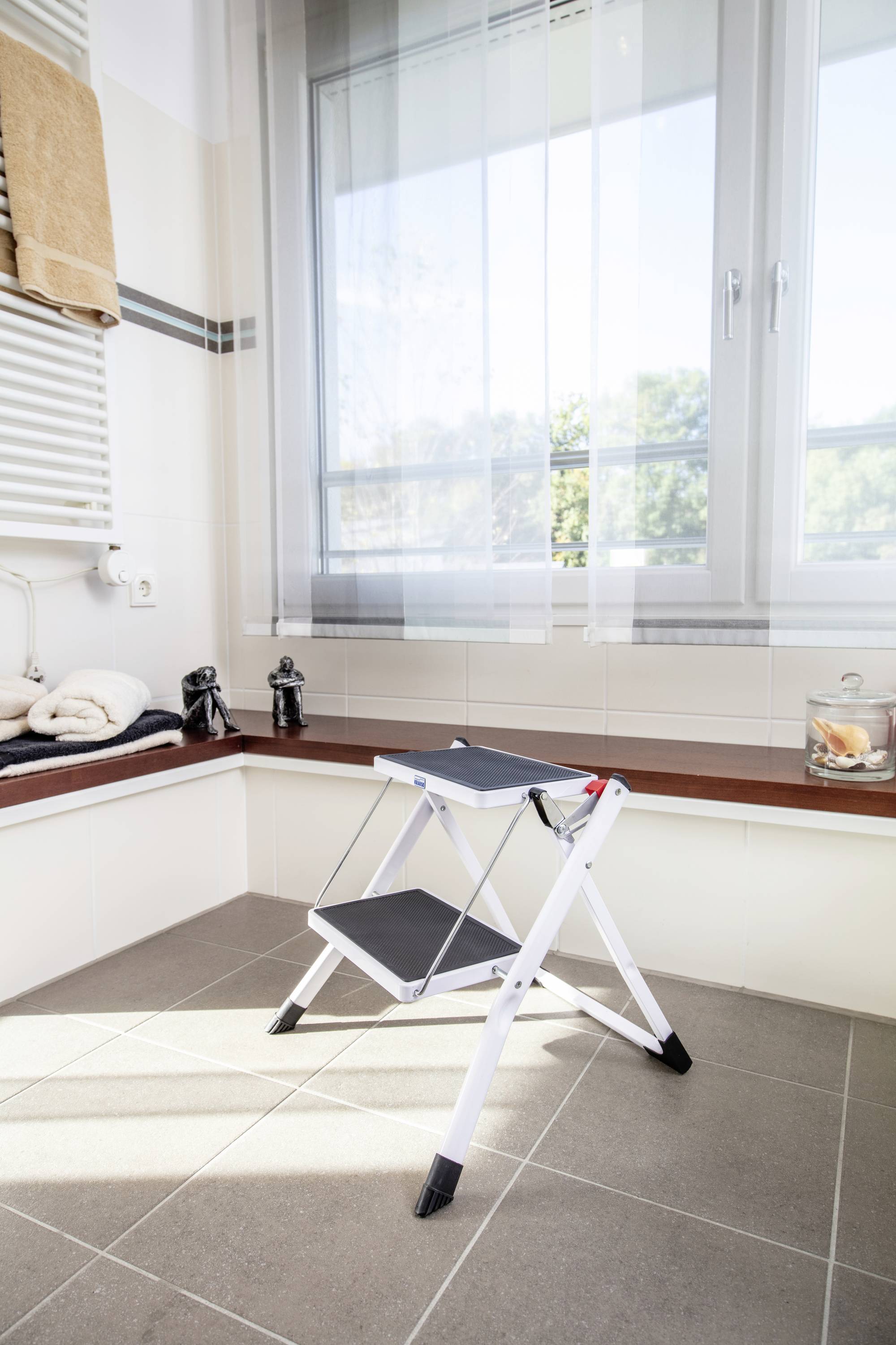 In einem hellen Badezimmer steht eine Klappleiter mit zwei Stufen aus Metall und Kunststoff auf dem gefliesten Boden vor einem großen Fenster.
