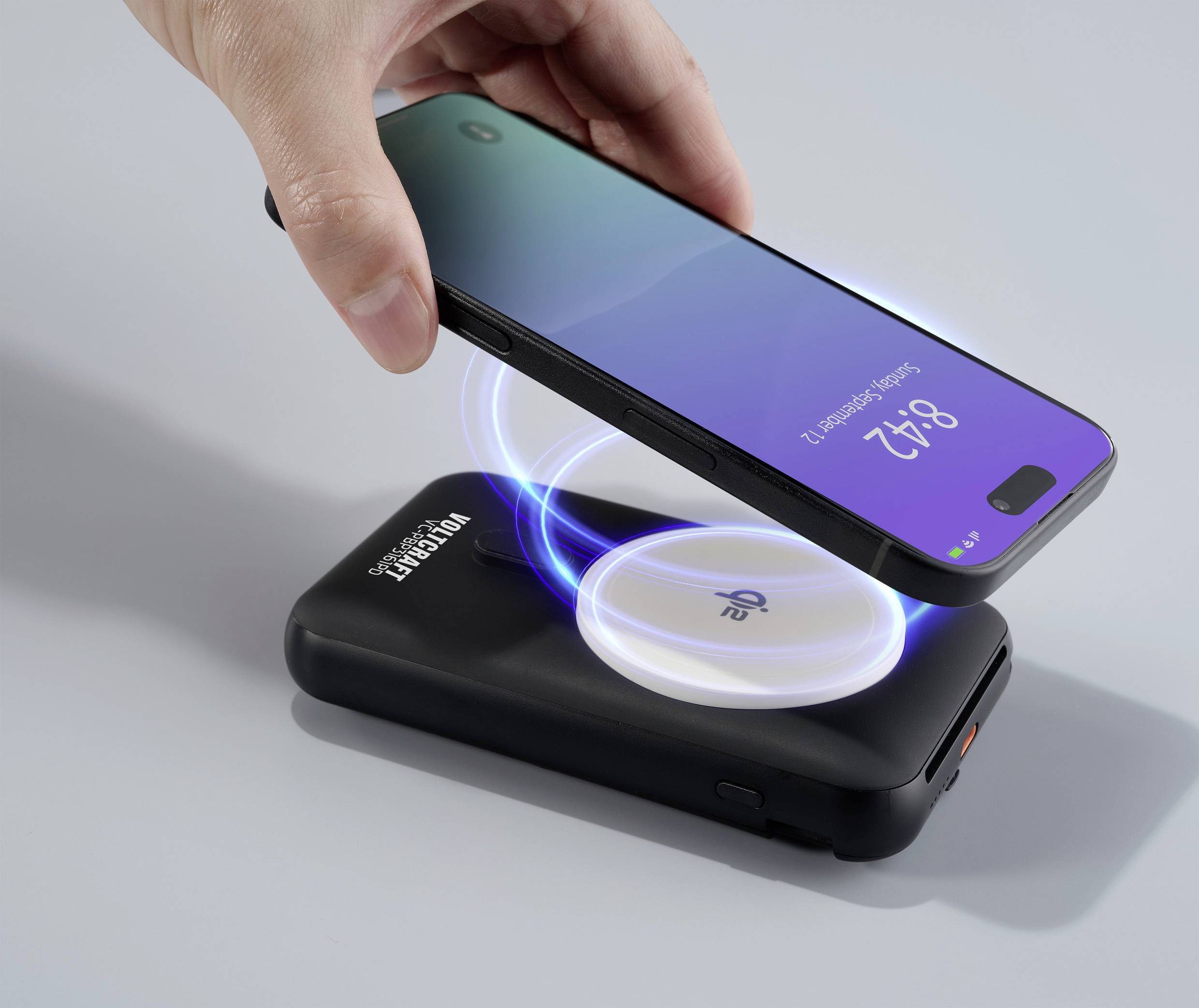 Ein Smartphone wird kabellos auf einem Ladegerät aufgeladen. Das Display zeigt die Uhrzeit 12:16 an. Hände halten das Smartphone.