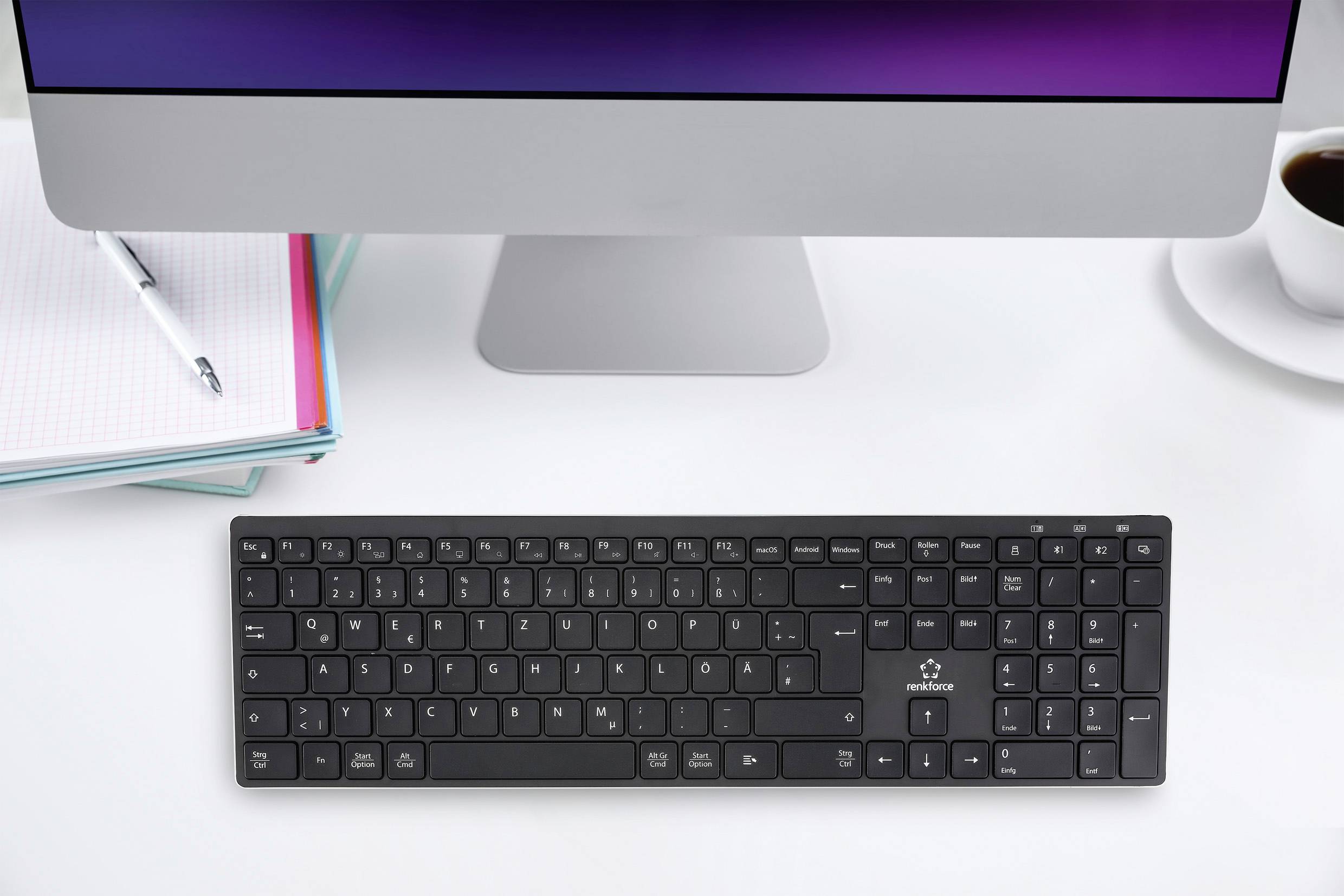 Schwarze Tastatur auf einem weißen Schreibtisch vor einem Computer-Monitor, daneben ein Notizbuch und eine Tasse Kaffee.