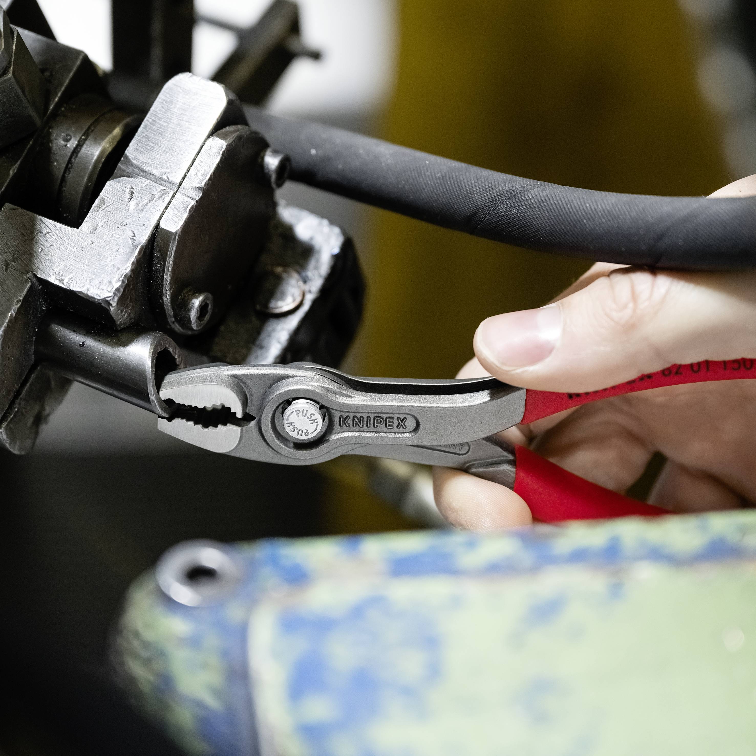 'Eine Hand hält eine Zange und zieht an einem schwarzen Kabel, das in einer Metallhalterung fixiert ist.'