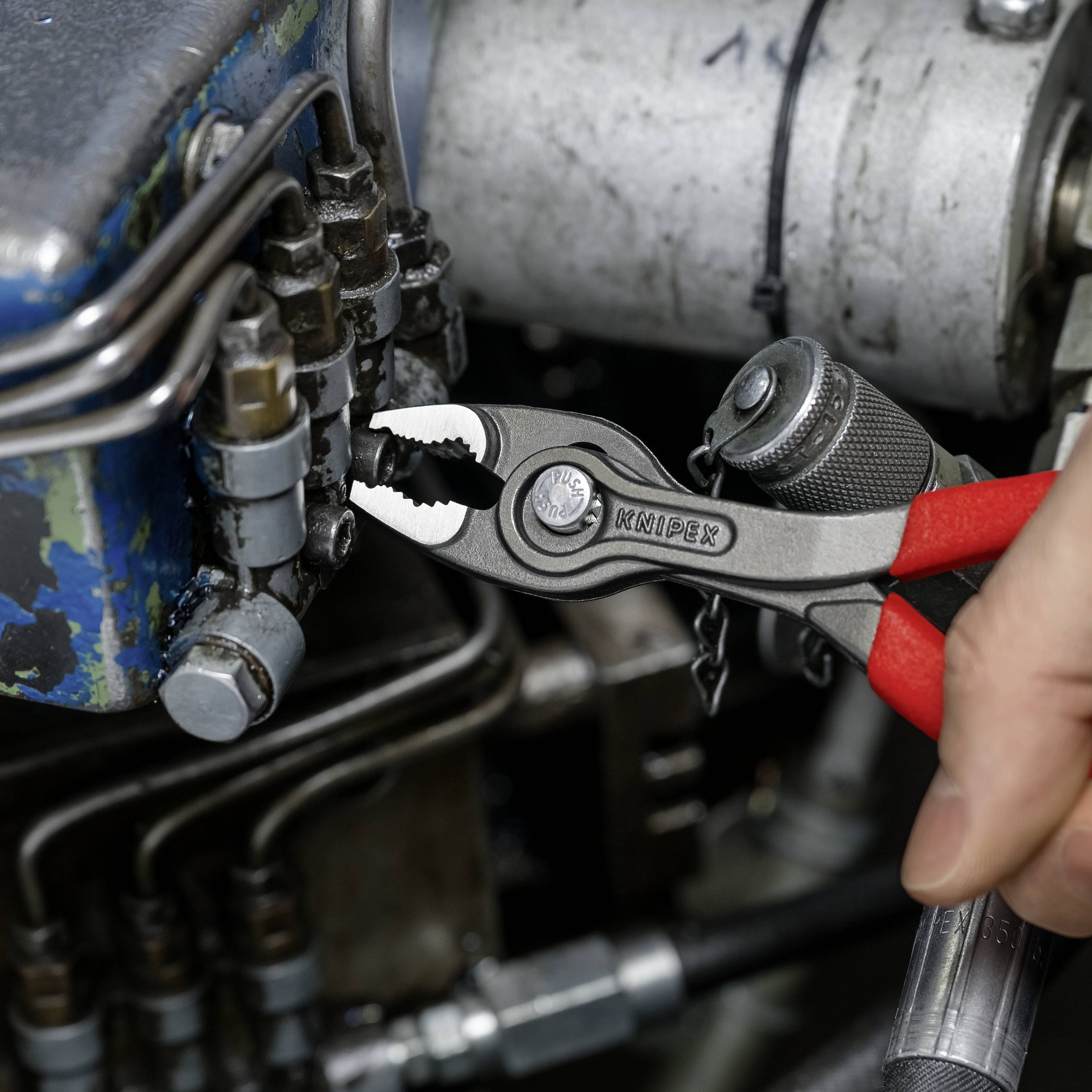 Eine Person zieht die Schraube einer alten, blauen Maschine mit einer Zange fest. Im Hintergrund sind Motorenteile zu sehen.