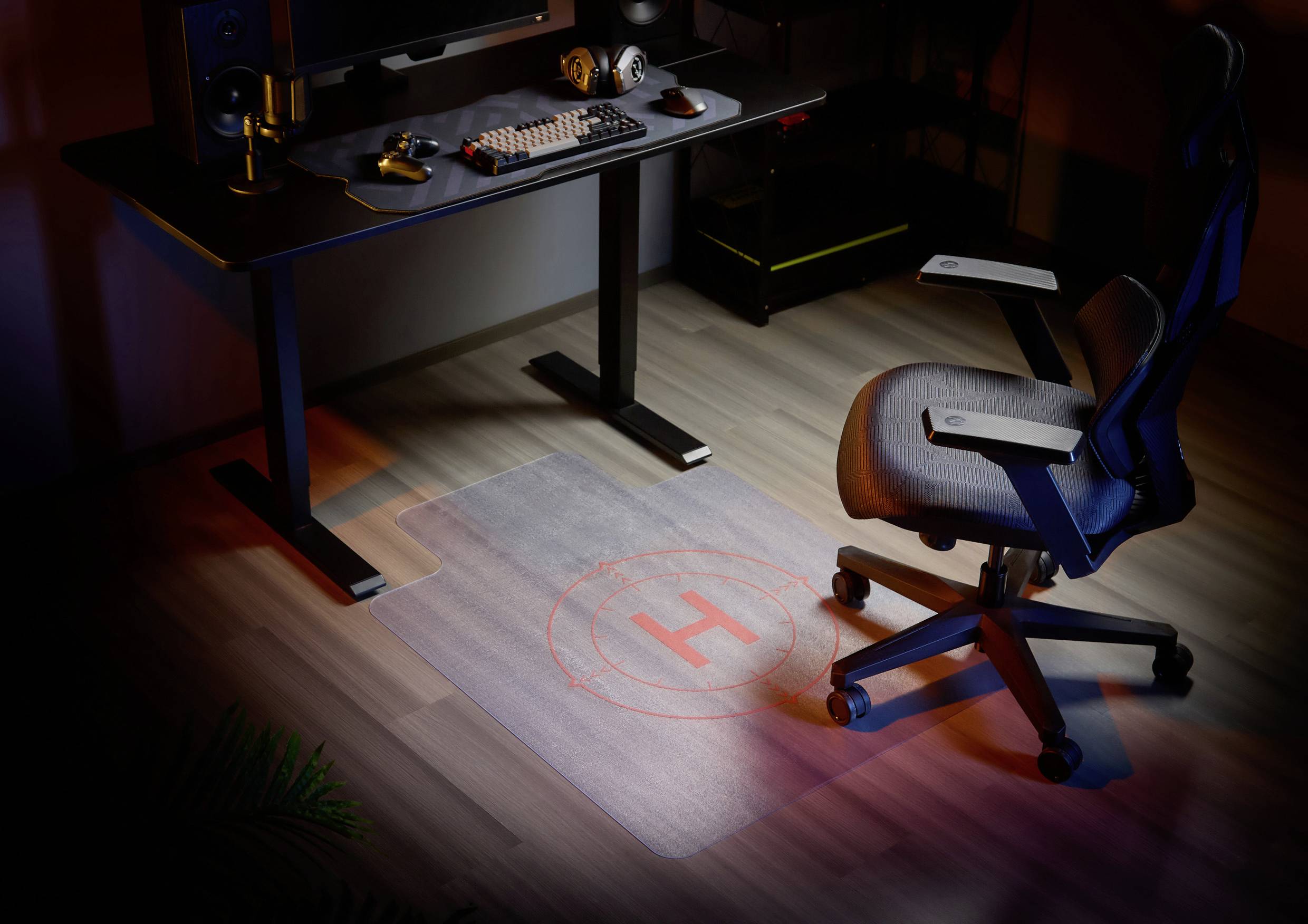 Ein Büro mit einem Schreibtisch, der Computer, Kopfhörer und Tastatur trägt. Auf dem Boden liegt eine transparente Matte mit einem roten 'H'.