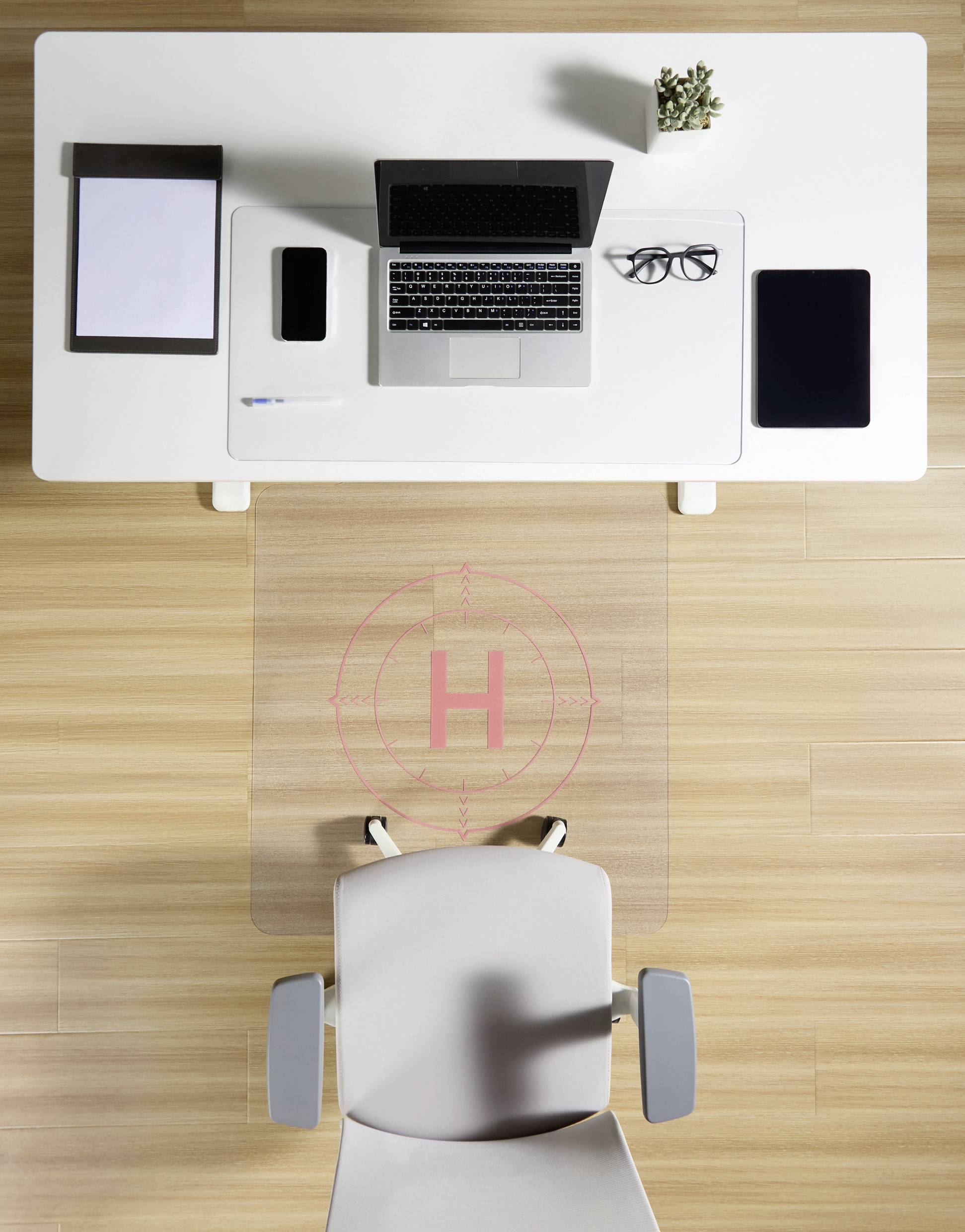 Ein moderner, aufgeräumter Schreibtisch mit Laptop, Smartphone, Brille und Tablet, darunter ein Bürostuhl auf einer durchsichtigen Matte.