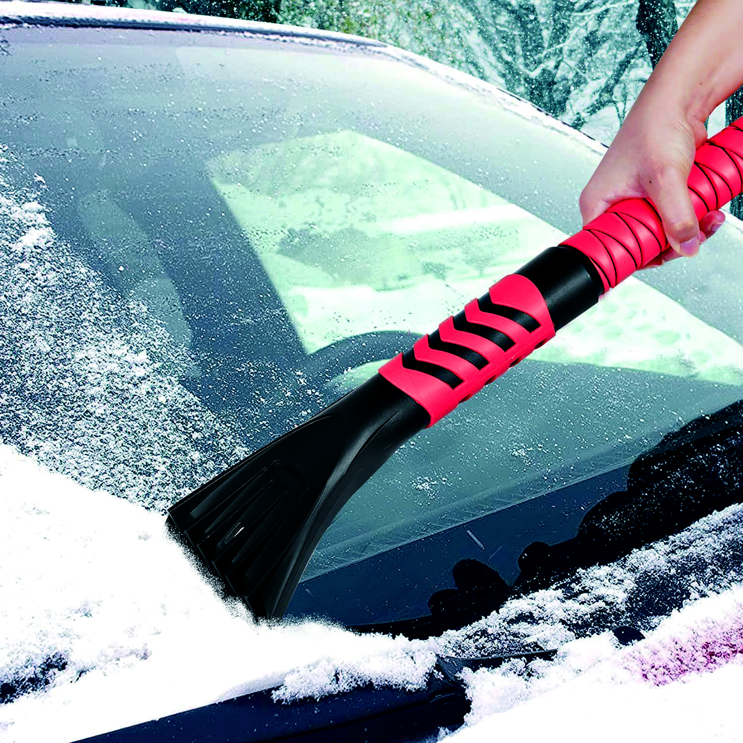Eine Hand benutzt einen Eiskratzer mit rotem Griff, um Schnee und Eis von der Windschutzscheibe eines Autos zu entfernen.