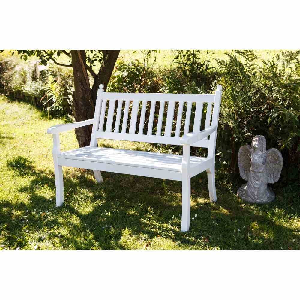 Weiße Gartenbank unter einem Baum im Sonnenschein, umgeben von Gras und Pflanzen. Rechts neben der Bank steht eine kleine Engelstatue.