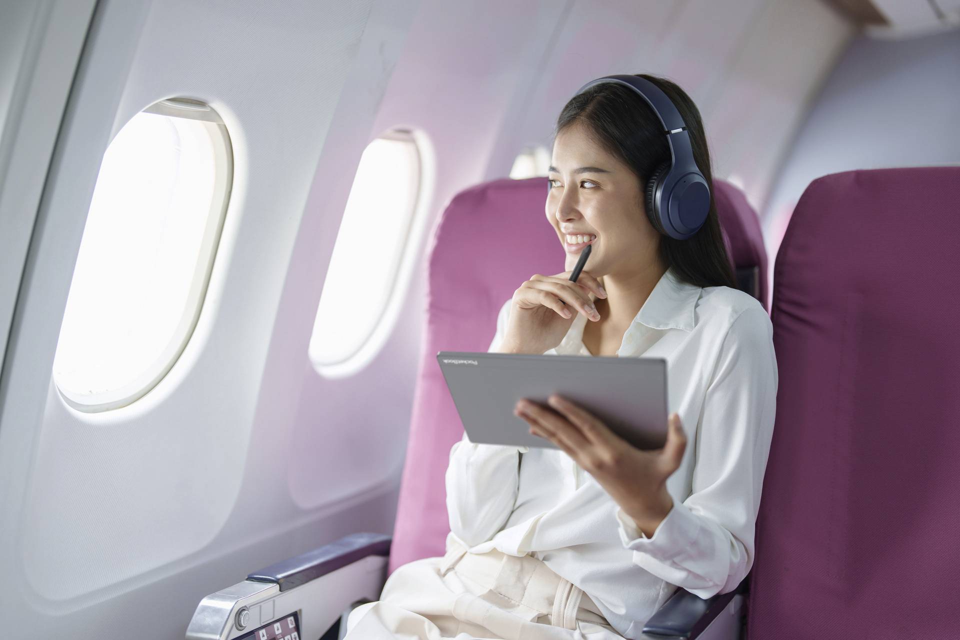Frau mit Kopfhörern lächelt und hält ein Tablet in der Hand, während sie in einem Flugzeugsitz am Fenster sitzt.