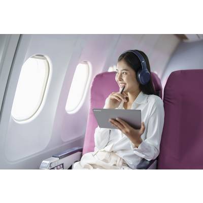Frau mit Kopfhörern lächelt und hält ein Tablet in der Hand, während sie in einem Flugzeugsitz am Fenster sitzt.