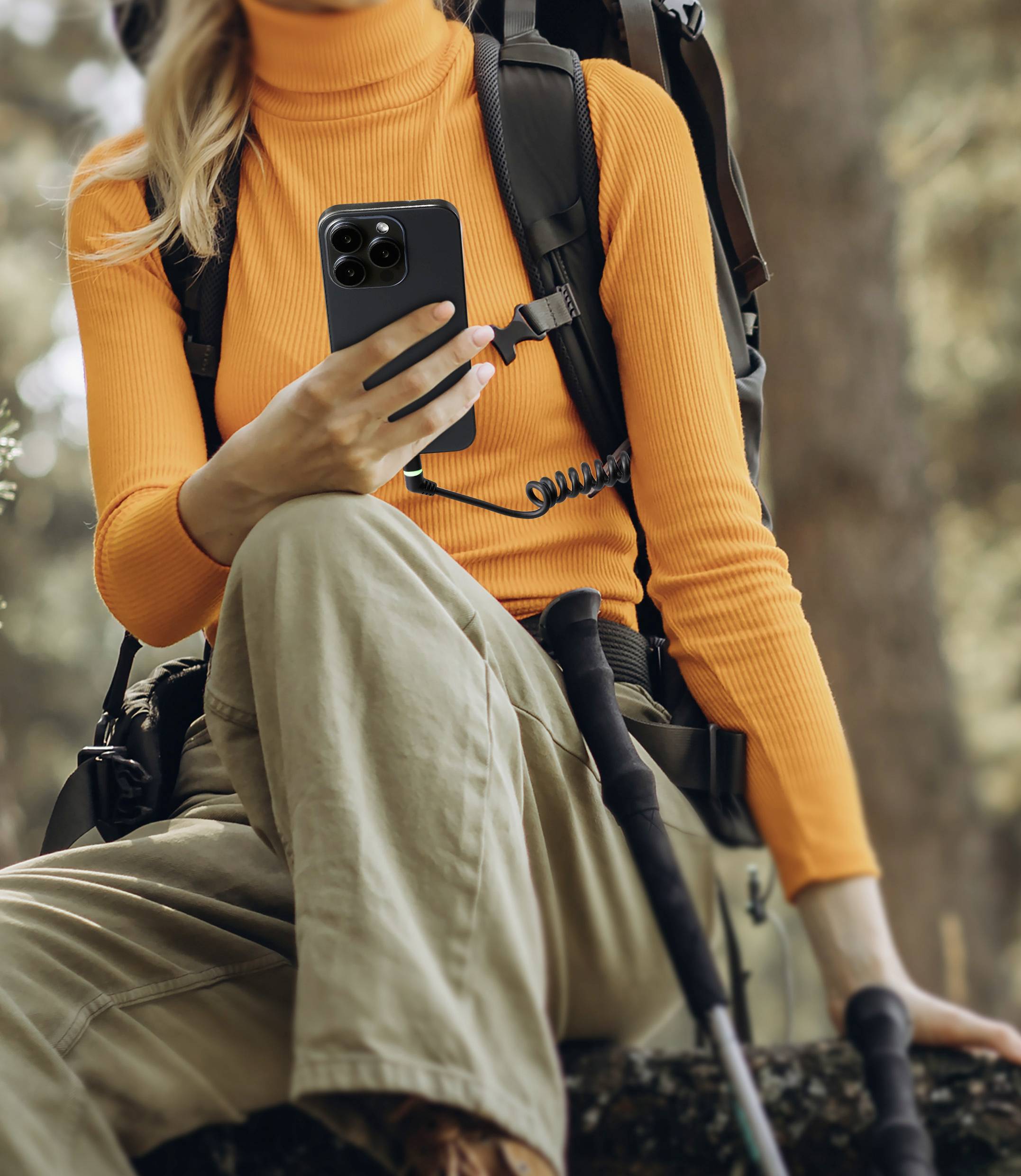 Eine Person im orangefarbenen Pullover sitzt im Freien mit einem Rucksack auf dem Rücken und schaut auf ein Smartphone.
