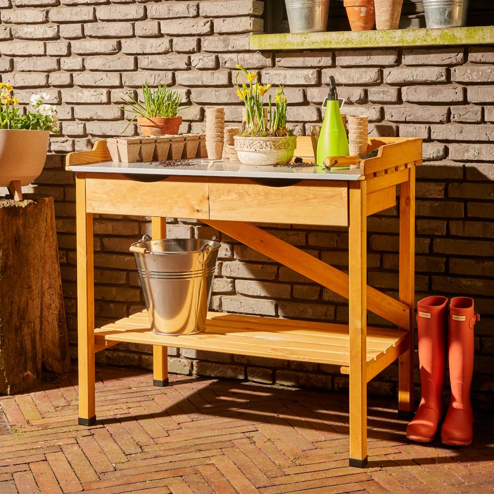 Holztisch im Garten mit einem Eimer, Gießkanne, Töpfen und blühenden Pflanzen. Im Hintergrund eine Ziegelmauer und der Boden ist aus Backstein.