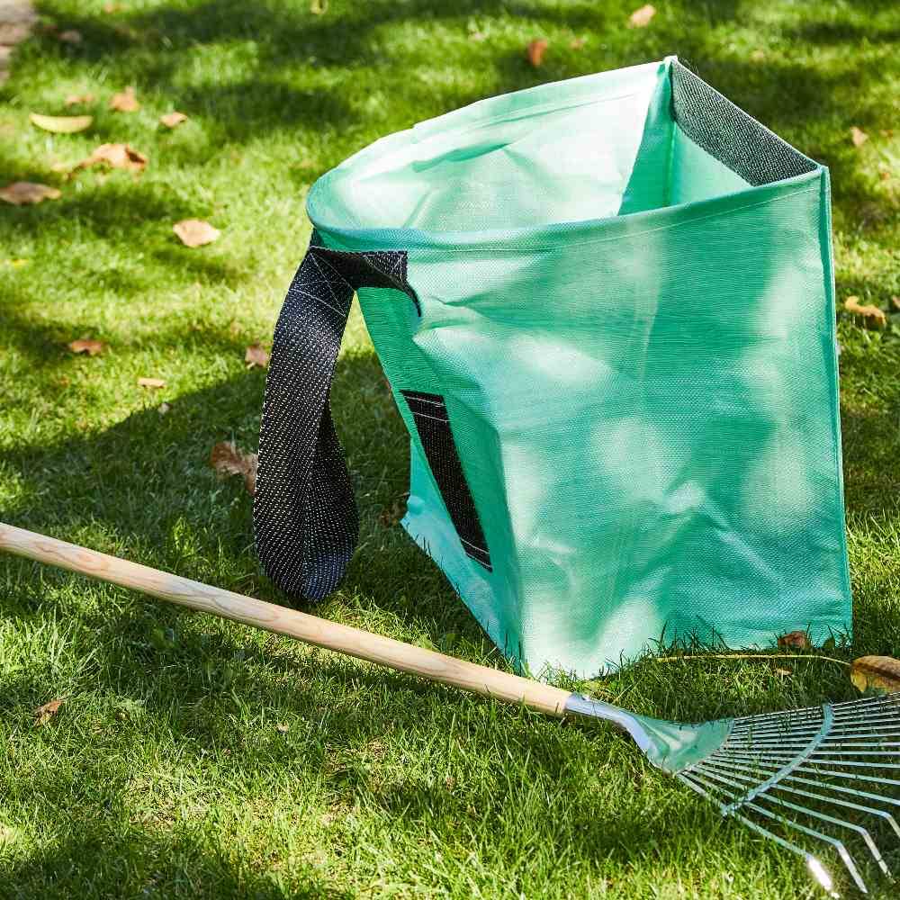 Grüner Gartenabfallsack steht auf Gras neben einem Rechen, umgeben von Laub. Scene zeigt Aufräumarbeiten im Herbst.