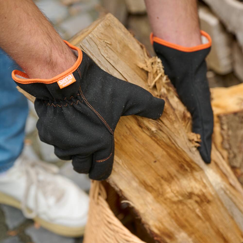 Eine Person trägt schwarze Handschuhe und hält ein Stück Holz. Im Hintergrund sind Holzkisten zu sehen.