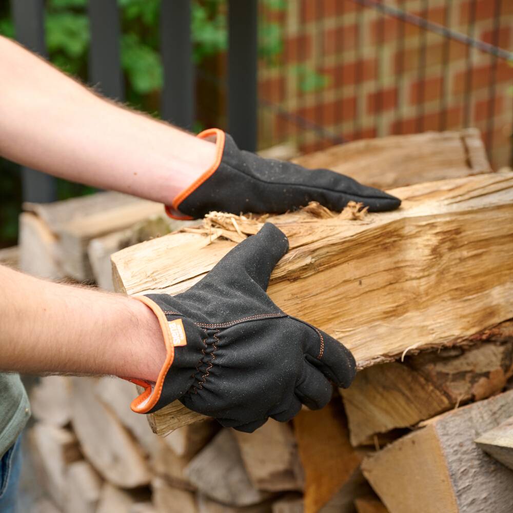 Eine Person mit schwarzen Handschuhen stapelt Holzscheite im Freien. Im Hintergrund sind weitere gestapelte Holzscheite zu sehen.