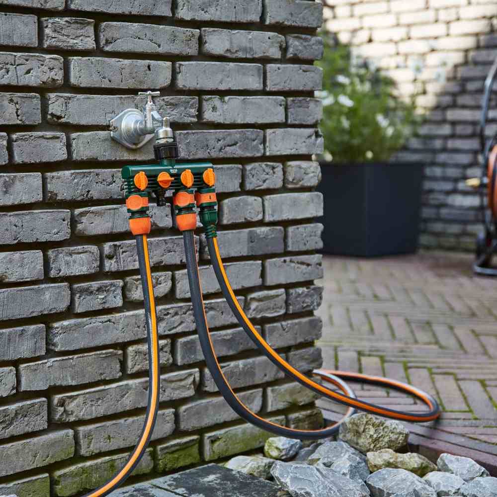 Ein Wasserhahn an einer Ziegelwand mit drei Gartenschläuchen, die an einen Verteiler angeschlossen sind, auf einem gepflasterten Hof.