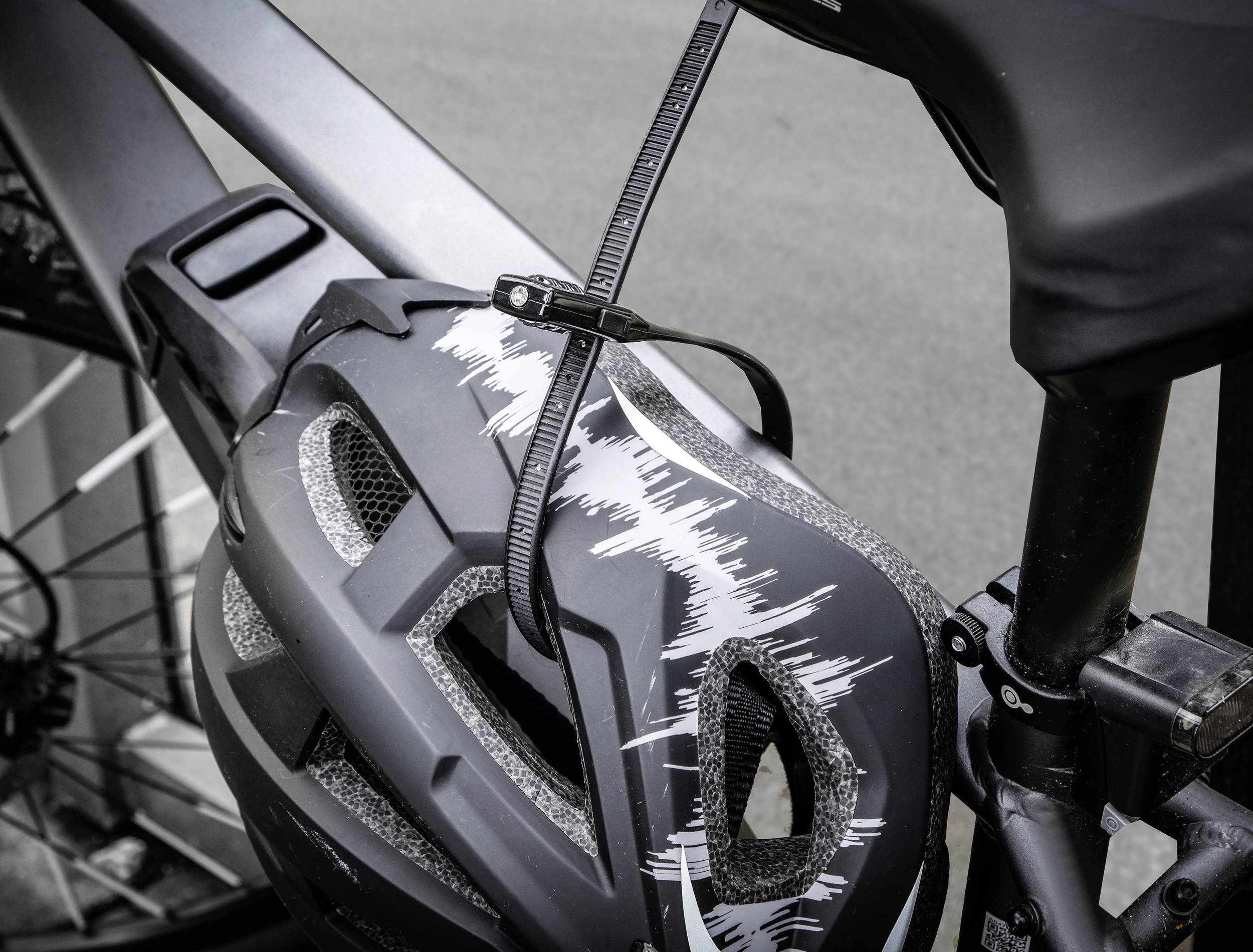 Ein Fahrradhelm ist mit einem Kabelbinder am Rahmen eines Fahrrads befestigt. Der Helm hat ein modernes Design mit grauen Akzenten.