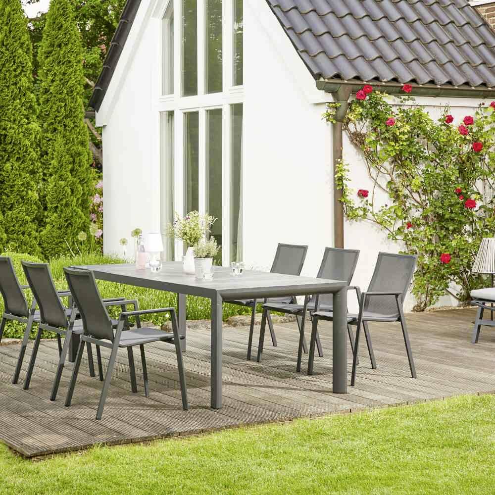 Ein moderner Garten mit einem grauen Tisch und sechs Stühlen auf einer Holzterrasse neben einem weißen Haus mit großen Fenstern.