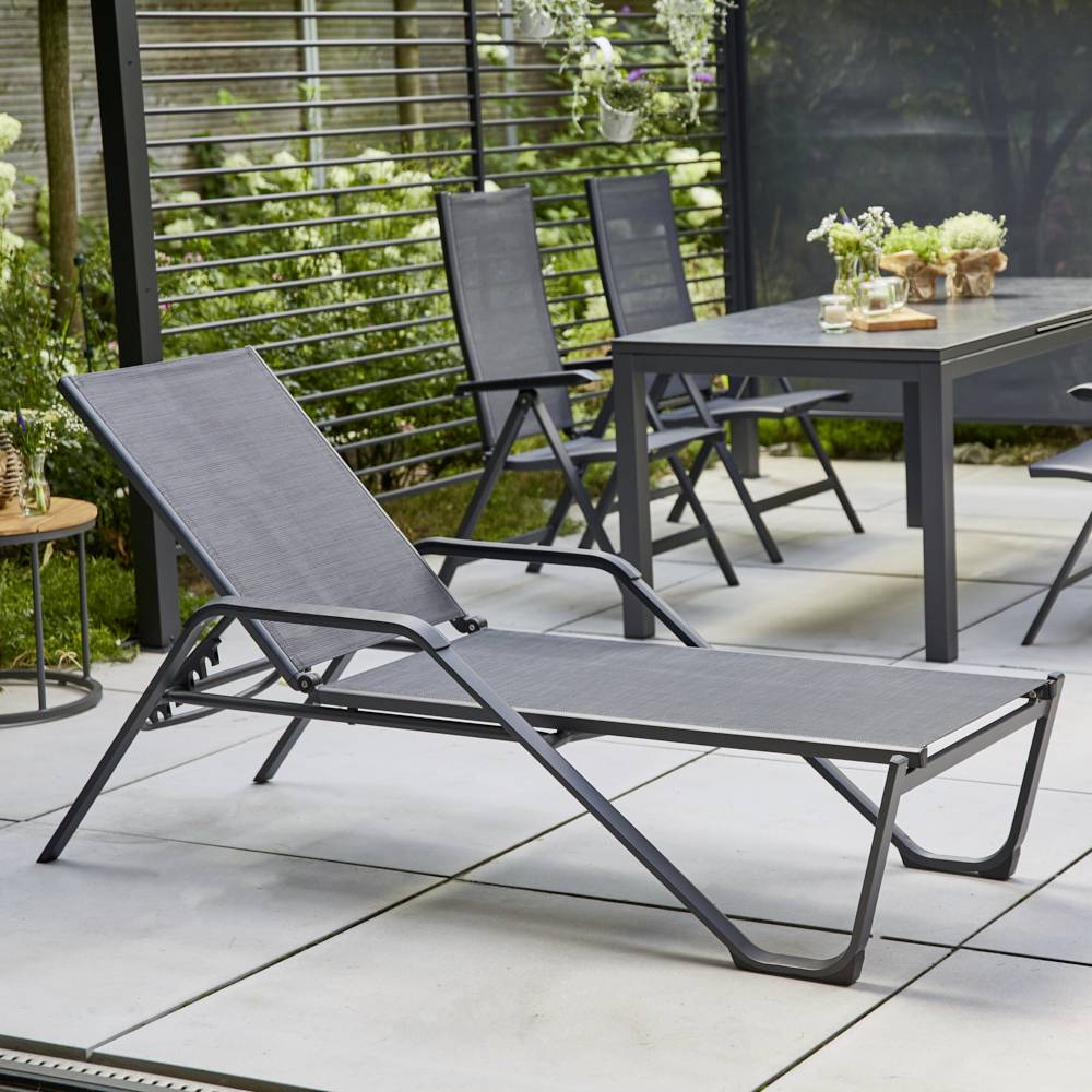 Liegestuhl auf einer Terrasse neben einem Gartentisch mit Stühlen, umgeben von Pflanzen, zeigt eine gemütliche Außensitzgelegenheit.