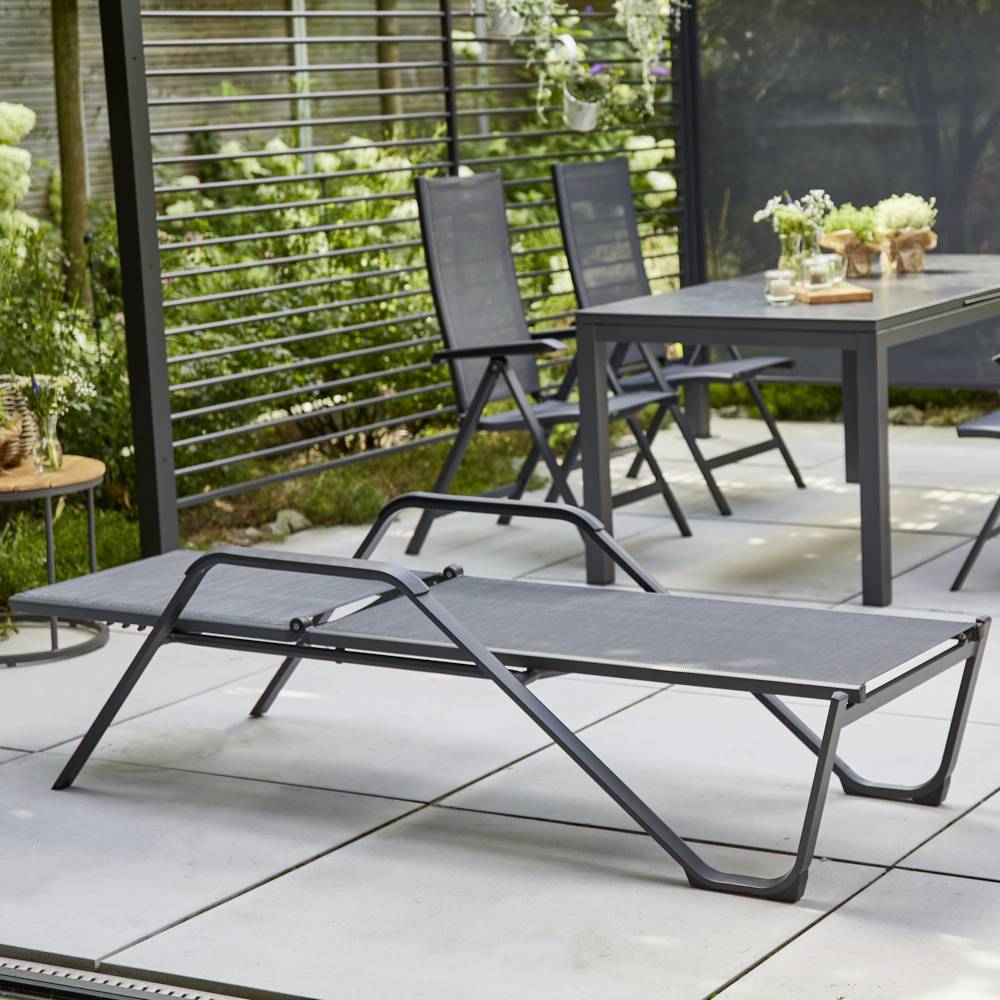 Garten mit grauer Sonnenliege, Esstisch und Stühlen auf Terrassenfliesen, umgeben von Pflanzen. Ruhige Outdoor-Entspannung.