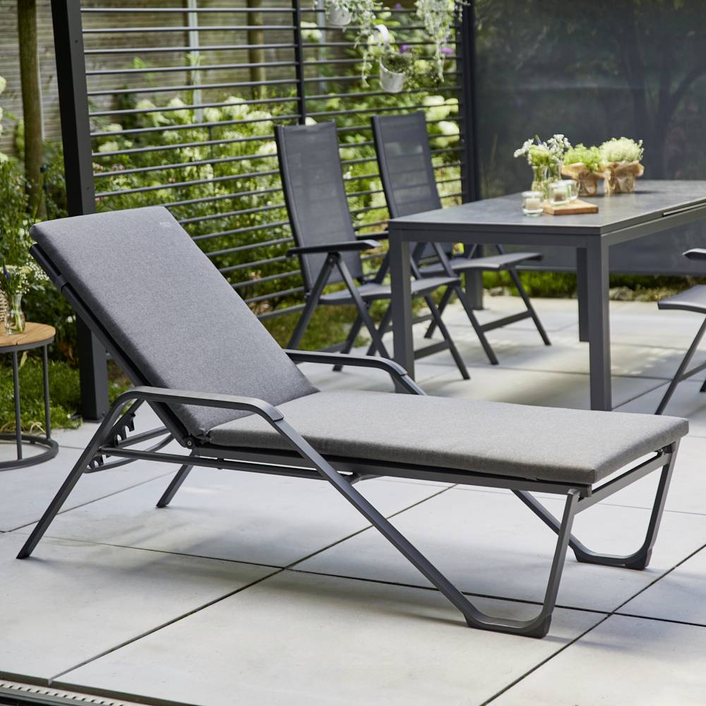 Garten mit moderner grauer Liege im Vordergrund, umgeben von einem Tisch und vier Stühlen. Pflanzen und Sichtschutz im Hintergrund.