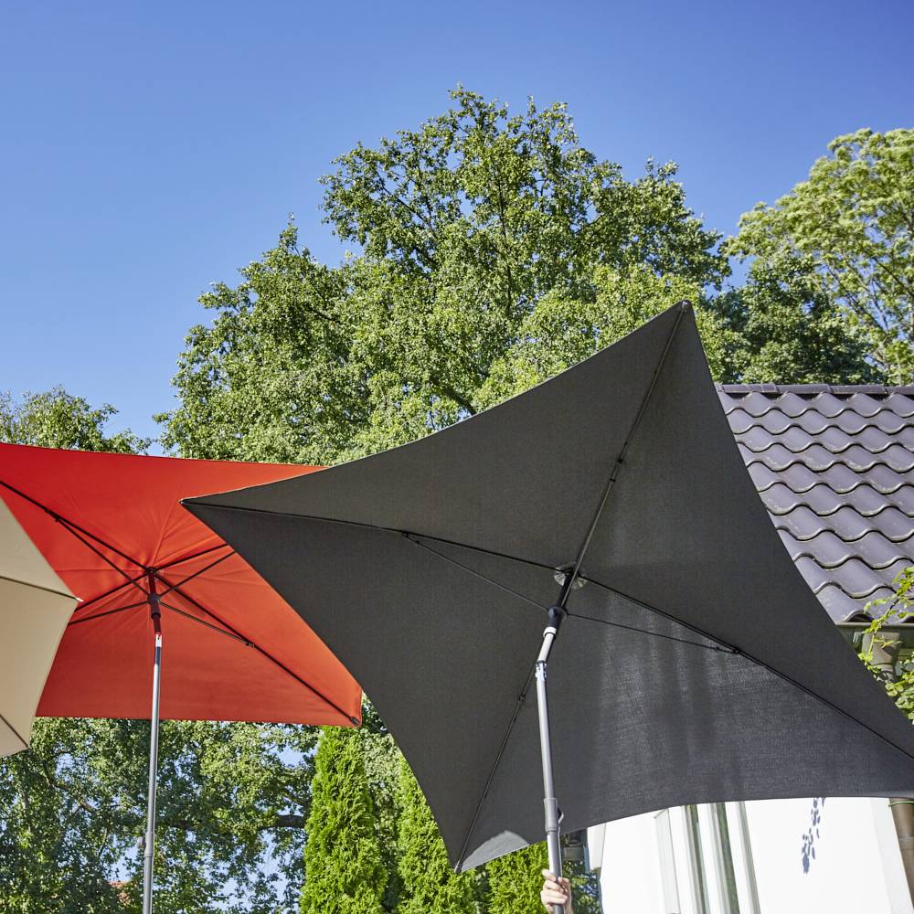 Zwei große, schattenspendende Sonnenschirme, einer rot, einer grau, im Garten bei Sonnenschein. Im Hintergrund grüne Bäume und ein Hausdach.