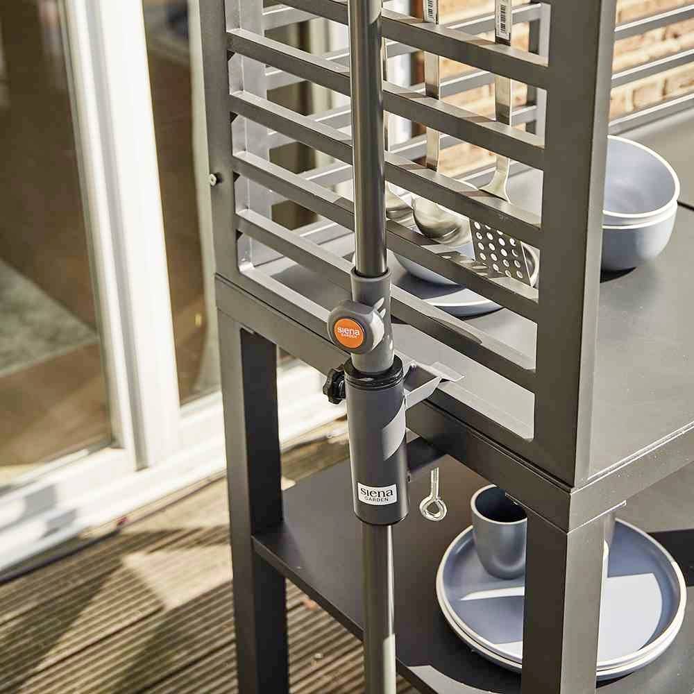 Ein schwarzer Metalltisch mit integriertem Grillutensilienhalter auf einer Terrasse. Auf dem Tisch steht blaues Geschirr.