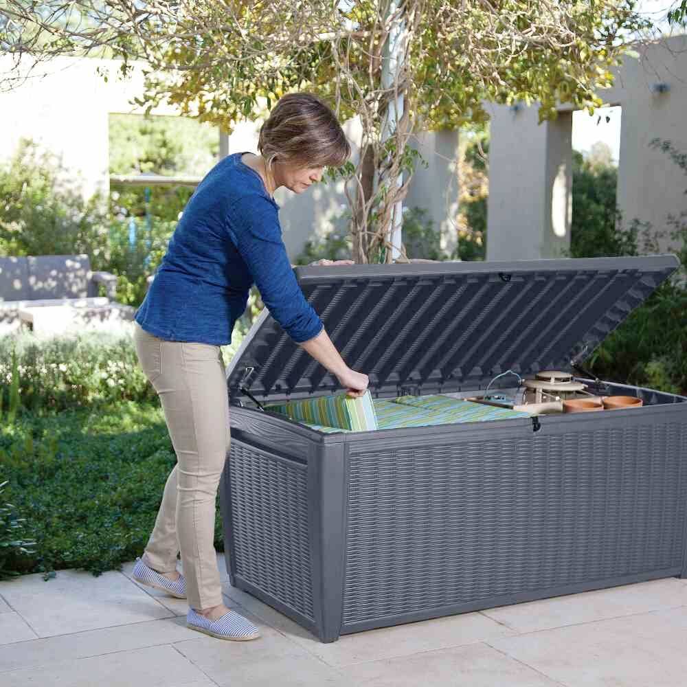 Eine Frau steht im Garten und räumt Gegenstände in eine große, offene graue Aufbewahrungsbox. Bäume und Sträucher im Hintergrund.