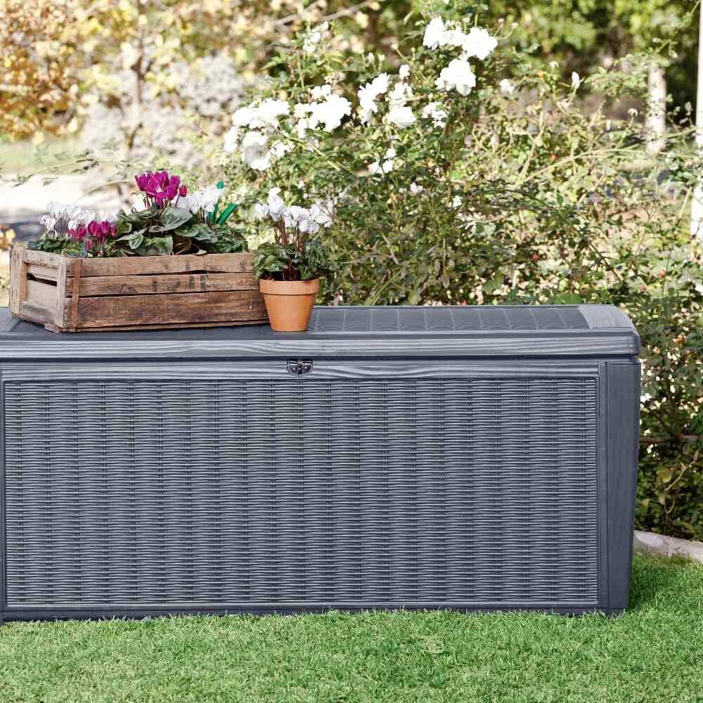 Große graue Aufbewahrungsbox im Garten, darauf ein Blumenkasten mit bunten Blüten und ein kleinerer Topf, umgeben von Pflanzen.