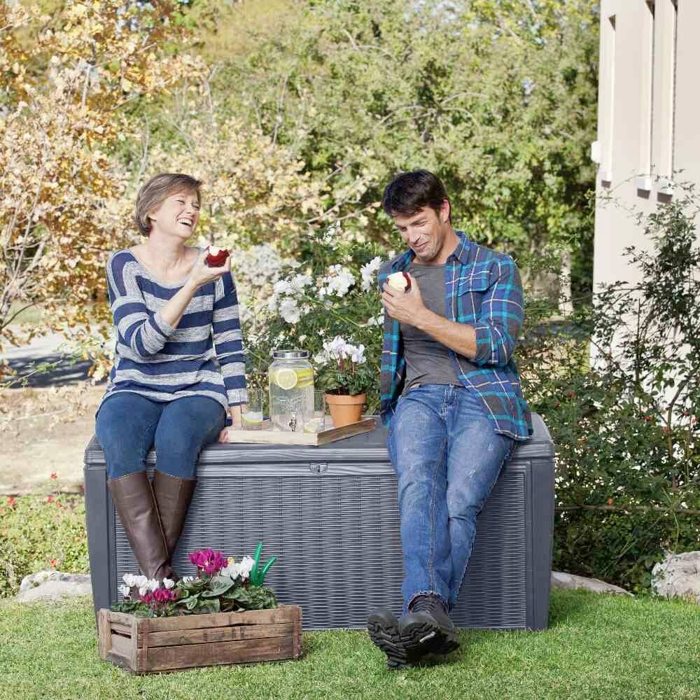Zwei lachende Personen sitzen auf einer grauen Gartenbox und essen Äpfel. Eine Holzkiste mit Blumen steht daneben, im Hintergrund blühende Pflanzen.