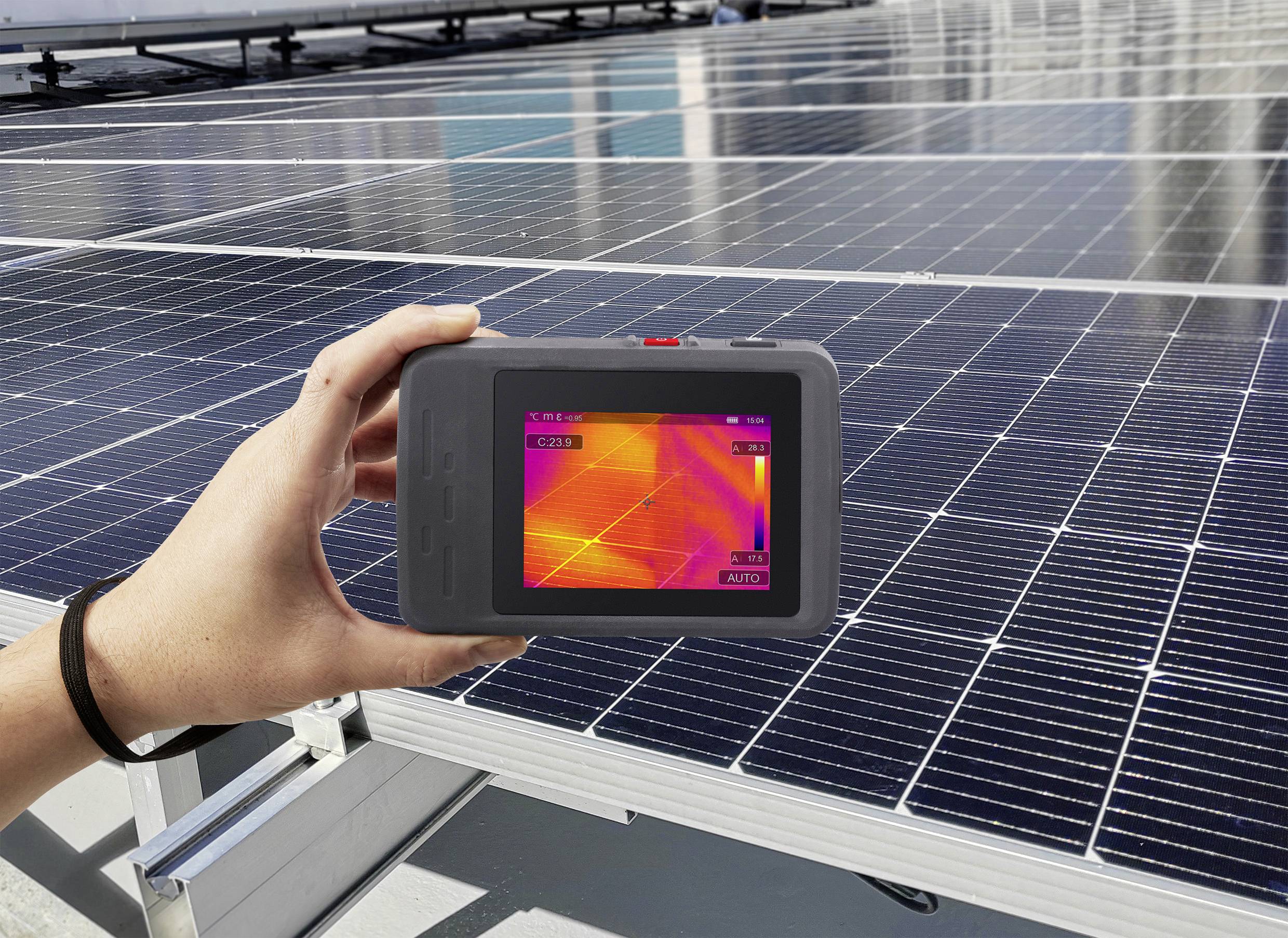 Hand hält Wärmebildkamera über Solarmodul, das Temperaturunterschiede zeigt. Hintergrund: weitere Solarmodule auf dem Dach.