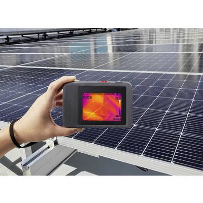 Hand hält Wärmebildkamera über Solarmodul, das Temperaturunterschiede zeigt. Hintergrund: weitere Solarmodule auf dem Dach.