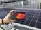 Hand hält Wärmebildkamera über Solarmodul, das Temperaturunterschiede zeigt. Hintergrund: weitere Solarmodule auf dem Dach.
