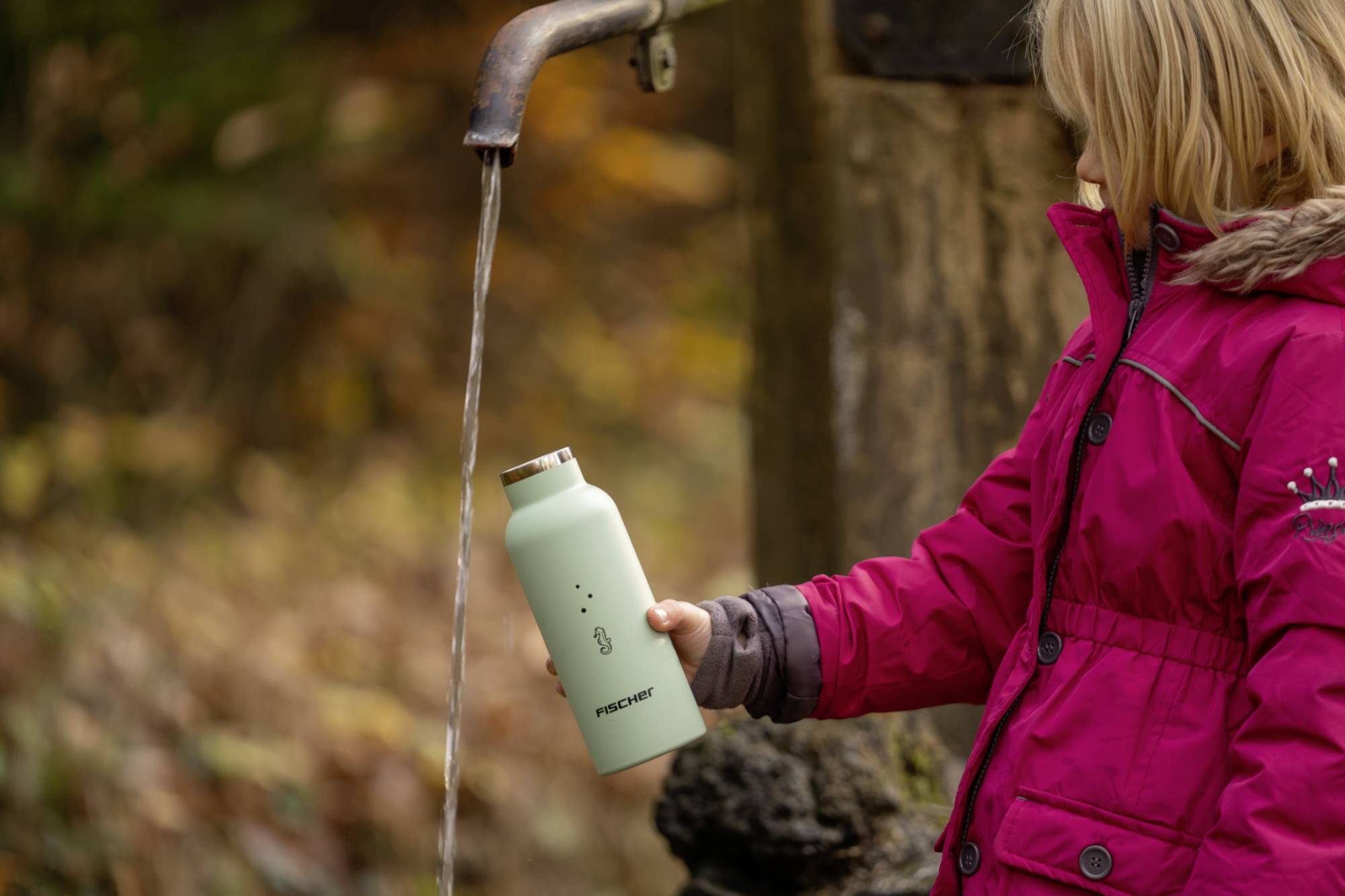 Ein Kind in einem roten Mantel hält eine Flasche unter einen Wasserhahn im Freien, um sie mit Wasser zu füllen.