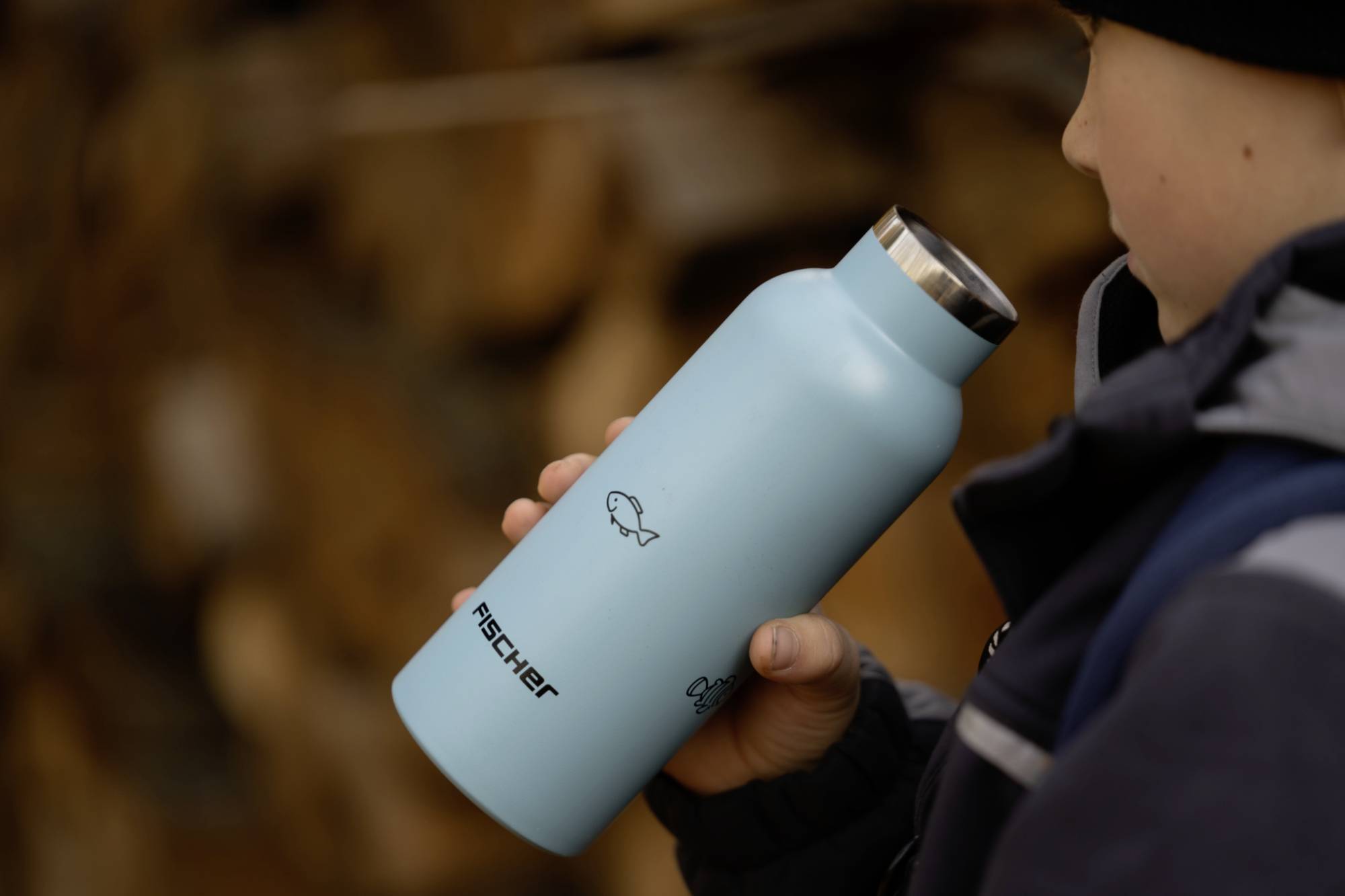 Ein Kind hält eine blaue Trinkflasche mit Fischmotiv und dem Schriftzug 'Fischer'. Die Flasche hat einen silbernen Verschluss.
