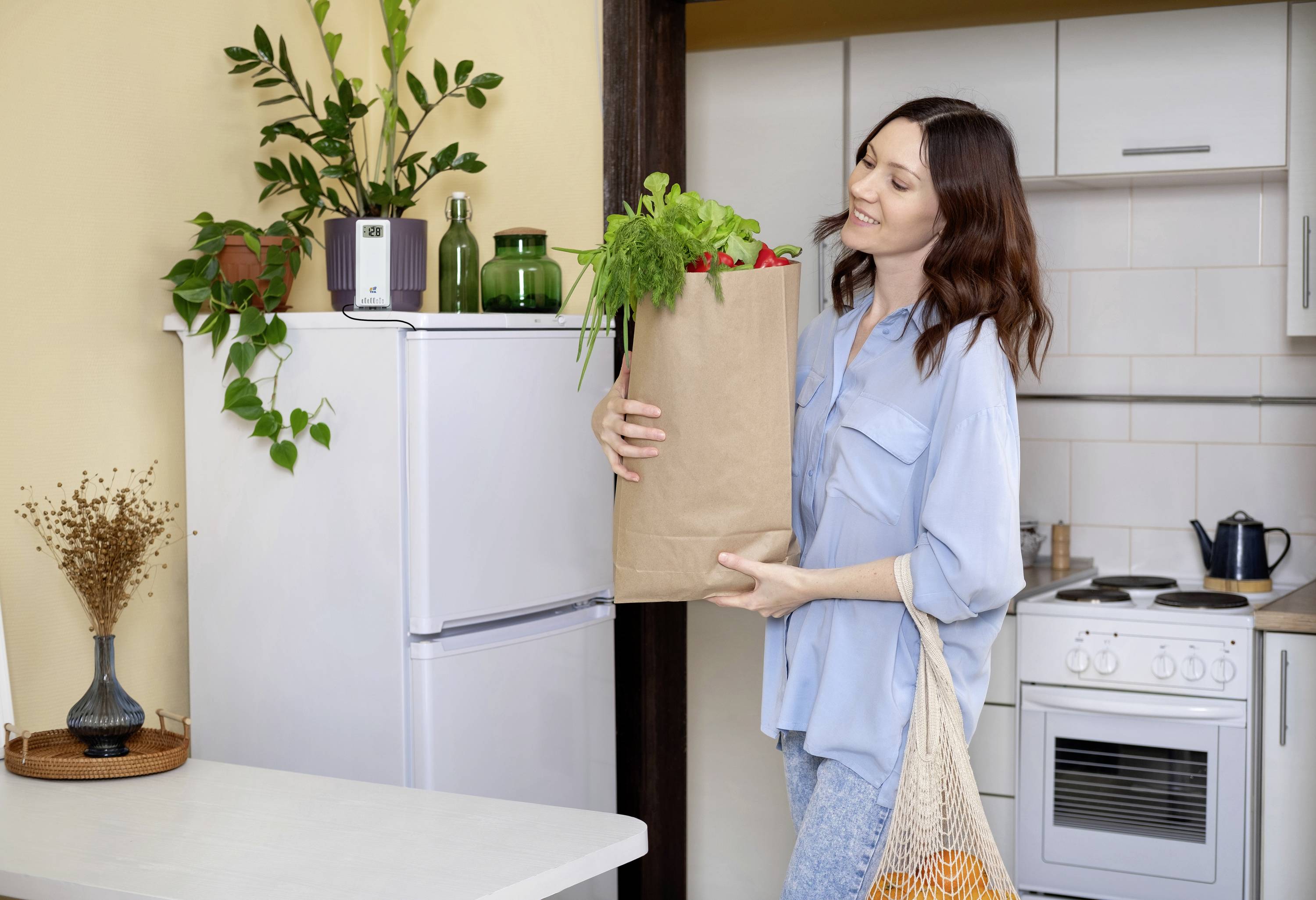 Eine Frau räumt eine Papiertüte voller frischem Gemüse in eine helle, moderne Küche ein. Im Hintergrund stehen Pflanzen auf einem Kühlschrank.