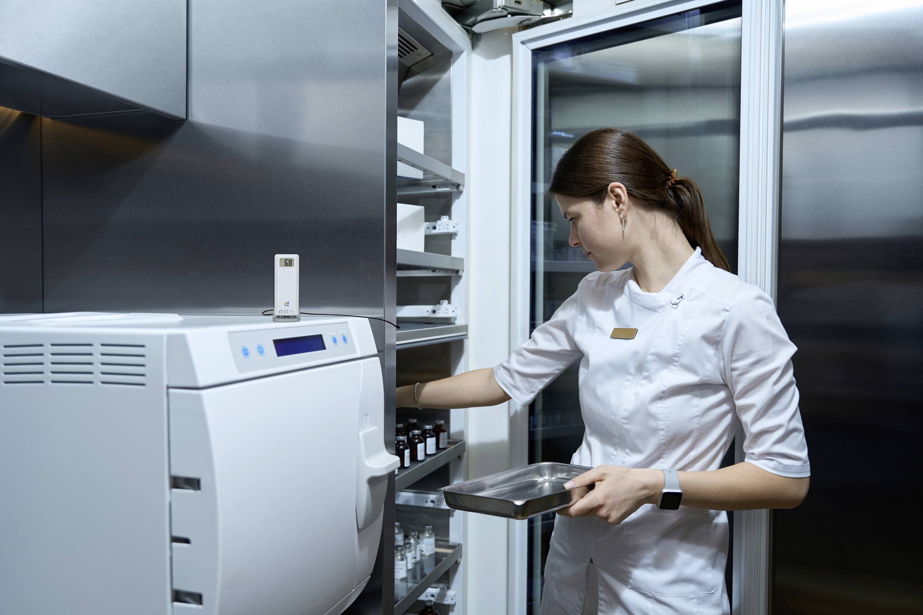 Eine Frau in weißer Berufskleidung entnimmt ein Medikament aus einem Kühlschrank in einem medizinischen oder pharmazeutischen Umfeld.