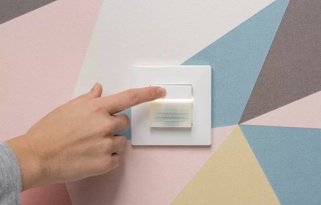 Eine Hand schaltet einen Lichtschalter ein. Der Hintergrund zeigt eine Wand mit geometrischen Mustern in Pastelltönen.