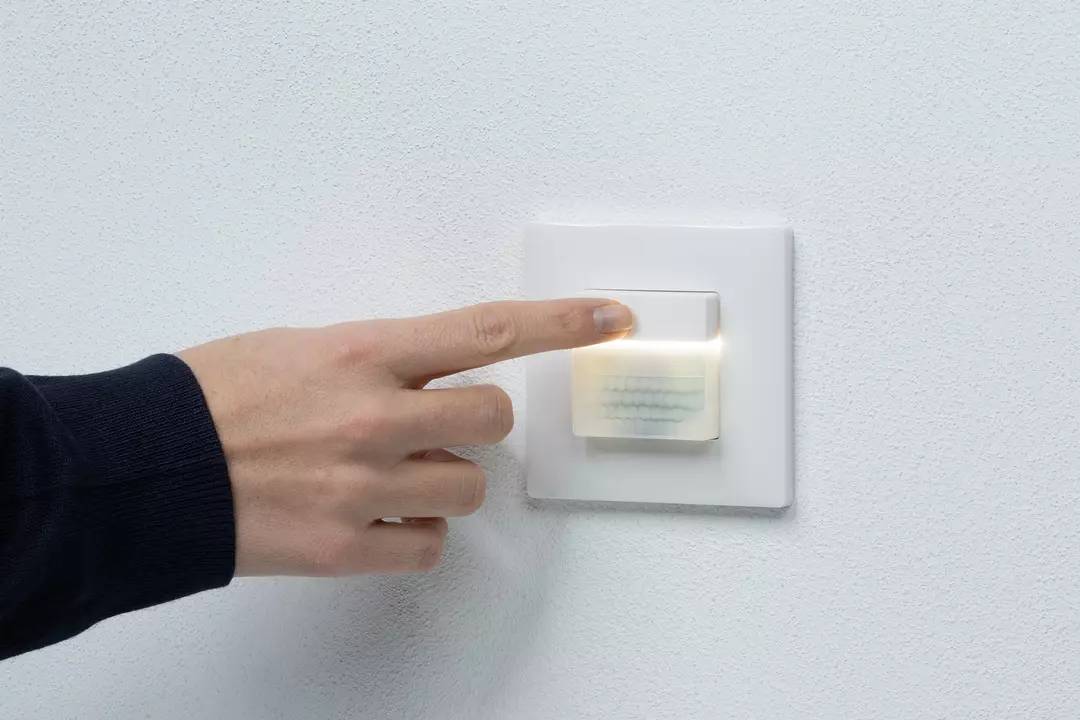Eine Hand drückt auf einen beleuchteten Lichtschalter an einer weißen Wand.