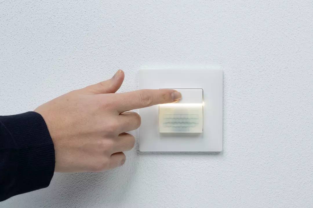 Eine Hand drückt einen beleuchteten Lichtschalter an einer weißen Wand.